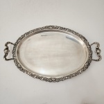 Belo tabuleiro oval em metal prateado, manufatura Meridional  E.P.S, apresentando rica borda trabalhada com ornamentação em relevo de inspiração rococó, composta por volutas, concheados e folhagens entrelaçadas. Alças laterais vazadas, de desenho sinuoso e equilibrado, integrando-se harmonicamente ao conjunto. Superfície ampla e lisa, ideal para serviço ou composição decorativa. Peça de elegante presença e excelente estado geral, representativa da produção brasileira de prateados da primeira metade do século XX. Dimensões: 53 x 34 centímetros.