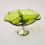 Elegante compoteira em vidro artístico verde, de forma orgânica suavemente ondulada, apoiada sobre base em prata esterlina (Sterling 925) da manufatura americana Empire, modelo 389, com núcleo weighted. A taça superior apresenta divisórias triangulares integradas ao próprio vidro, criando desenho moderno e funcional, enquanto o pedestal em prata confere equilíbrio, brilho e sofisticação ao conjunto. Peça decorativa de forte apelo estético, representativa do diálogo entre vidro mid-century e prataria americana do século XX. Dimensões: 13 x 20 centímetros.