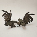 Par de esculturas decorativas representando galos em combate, confeccionadas em metal banhado a prata, com detalhamento minucioso de plumagens, asas abertas e caudas elevadas que conferem grande dinamismo e teatralidade à composição. O acabamento em prata realça os volumes e dá luminosidade à modelagem, mantendo pátina elegante. Conjunto típico da produção decorativa oriental do século XX. Dimensões: 20x22x18 centímetros. ( cada)