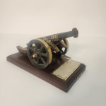 Elegante miniatura de canhão com estrutura metálica em acabamento escuro e detalhes dourados, apoiada sobre sólida base de madeira polida. Rica em detalhes, a peça evoca as tradições militares e o universo naval. Pertenceu a um almirante da Marinha Brasileira, acrescentando um contexto histórico discreto ao objeto.                                                       Estado de conservação: apresenta leves sinais do tempo e pequenas marcas de manuseio, mantendo excelente aparência geral. Medindo: 13x22x12