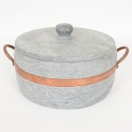 EQUIPAMENTO DE COZINHA, uma (1) panela com tampa, confeccionada em soapstone (pedra sabão), torneada,cinta e alça em metal cobreado, marca BRAS ART, medindo 26,5 cm diâmetro.