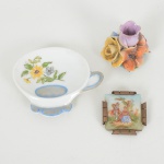 PORCELANA, três (3) itens: 1 suporte para saquinho de chá, formato de xícara, medindo 8 x 7 cm; 1 broche,ornado com cena de galanteio, possivelmente baseado na obra de FRAGONARD (Jean Honoré Fragonard, Grasse 1732Paris 1806, pintor, desenhista e gravurista francês, estilo Rococó, conhecido pela exuberância e hedonismo de sua obra,pinturas do gênero com atmosfera de intimidade e erotismo), entorno com faixa na tonalidade magenta, generososarremates a ouro, medindo 10 cm diâmetro e 7,5 cm altura; 1 na tonalidade azul cobalto, ornada com douração (falhasna douração), origem Limoges, França, monture em bronze, falta o alfinete, medindo 4 x 4 cm; 1 arranjo floralminiatura, policromado, origem Portugal, medindo 4 cm altura, vide foto.