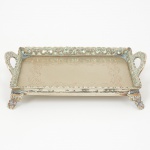 METAL PRATEADO, uma (1) pequena bandeja retangular, fundo burilado, borda vazada em volutas e elementosvegetalistas, pés com terminações em garra, medindo 17 x 12 cm, sinais de uso, vide foto.