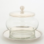 METAL PRATEADO E VIDRO, um (1) pote incolor e translúcido, tampa e presentoir em metal, borda relevadaem corda, pega da tampa esférica, punção não identificado no presentoir, medindo 10 cm altura, sinais de uso, vide foto.