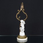 LUMINÁRIA, uma (1) antiga base para abat-jour para um lume, estrutura em bronze relevada em elementosvegetalistas, perdas na parte superior, restaurada com durepox, parte elétrica ausente, assente sobre mármore branco,contém escultura em biscuit na tonalidade branca, representando menino ouvindo uma concha marinha, medindo 35 cmaltura, vide foto.