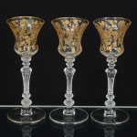 DEMI CRISTAL, três (3) taças de pé alto para licor, incolores e translúcidas, bojo ligeiramente canelado navertical, ornadas com elementos vegetalistas a ouro, fuste hexagonal na forma de balaustre com vários anéisintermediários em gomos e facetas, base em bolacha, um deles com bicado na base, medindo 12,5 cm altura, sinais deuso, vide foto.