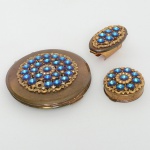 ACESSÓRIO FEMININO, conjunto com três (3) itens: 1 porta pílula; 1 porta batom com espelho acoplado; 1trousse com espelho interno, marca KIGU, Made in England, marcado em uma das peças, tampas com apliques emstrass azuis, faltando um deles, confeccionados em metal dourado, medindo a maior peça 8 cm diâmetro, sinais de uso,vide foto.