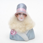 Caixa em porcelana half doll, início do século XX, esmaltada em tom azul-claro, adornada com delicada rosa modelada em relevo. A tampa é composta por uma elegante half doll em porcelana, dama de estilo Art Déco, com rosto finamente pintado, chapéu em tons de rosa e cinza e estola em tecido felpudo original. 12X8 cm medida total da caixa com a tampa