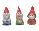 Conjunto de três pequenos gnomos decorativos em porcelana policromada, modelados e pintados à mão, cada figura com chapéu pontudo vermelho e características faciais distintas. 7,5x5 cm (maior).