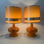 Par de abajures de cerâmica vitrificada, apresentando bases bojudas em tom alaranjado, com recortes vazados de inspiração geométrica e floral, conferindo efeito decorativo quando iluminados.  Acompanha cúpulas originais de época, em tom claro com detalhes de faixas largas metálicas horizontal. Medida total 50x28 cm com a cúpula