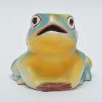 Escultura decorativa representando sapo em cerâmica, com olhos salientes e boca aberta. Acabamento esmaltado com policromia em tons de verde, amarelo e nuances terrosas. Possui pequeno fio de cabelo em sua pata esquerda.14x13 cm.