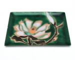 Travessa de metal inteiramente esmaltado, decorada com flor estilizada em tons de branco, verde e dourado sobre fundo verde profundo, esmalte de acabamento brilhante, O verso é esmaltado em tom azul-escuro uniforme. 21x17 cm