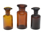 Conjunto composto por três frascos em vidro, de uso farmacêutico ou laboratorial, em tonalidades: âmbar claro, âmbar escuro e violeta-escuro (quase negro). Corpos cilíndricos, com base plana e gargalo curto, apresentando boca larga com rebordo reforçado, própria para vedação com rolha ou tampa de vidro esmerilhado. Alturas 11 e 9,5 cm