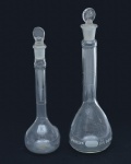 Par de balões volumétricos em vidro incolor, de corpo globular e pescoço longo e estreito, com tampas esmerilhadas originais em vidro. Apresentam marcações de capacidade gravadas a ácido, visíveis na parte inferior dos corpos: um exemplar com capacidade de 50 ml e outro de 25 ml. Peças de precisão destinadas a uso laboratorial ou farmacêutico. 15 e 13 cm altura