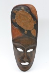 Máscara tribal em madeira entalhada, estilização facial alongada com olhos vazados e boca recortada, decorada com motivo floral em relevo. 39x16 cm.