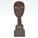 Escultura em madeira maciça, de linguagem popular, representando cabeça humana estilizada. O rosto apresenta olhos amendoados com pintura contrastante, cílios incisos e nariz alongado em relevo, conferindo forte presença gráfica à peça. Peça de estética primitiva, muito adequada tanto para coleção quanto para ambientação. 28x8x8 cm