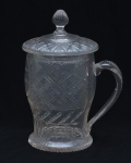 Caneca de grandes proporções destinada ao serviço de bebidas,  em vidro lapidado transparente, com decoração geométrica em padrão losangular e faixas de lapidação cruzada. Alça lateral robusta, moldada em vidro maciço. Acompanha tampa original em vidro, com lapidação radial e pegador em botão facetado, ajustada ao bordo superior. Datada de 1833. 23x17 cm
