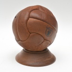 Réplica  - bola da copa do mundo 1950  em couro escuro, confeccionada em gomos costurados. Inscrições e marcas impressas, entre elas ''Almacén de Cueros'' e ''courroies veyrier''. Acompanha pedestal  em madeira torneada. Marcas do tempo. 19 cm diam (bola)  /  18x4 cm.