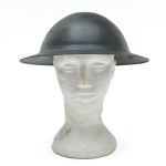 Capacete militar em aço, com aba curta e contínua, típico de modelos utilizados no século XX. Apresenta acabamento original, evidenciando desgaste natural, oxidação superficial e marcas de uso compatíveis com serviço ativo. Na parte frontal conserva insígnia metálica aplicada, composta por emblema central ladeado por estrelas, indicando provável uso oficial, associado a forças armadas. Interior desprovido do forro original, restando apenas elementos metálicos de fixação, comuns em capacetes de campanha. Possivelmente usado entre 1930 e 1950 (Segunda Guerra Mundial e anos imediatamente posteriores).