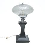 Anos 70 - Luminária de mesa de caráter escultórico de presença marcante e desenho singular, estruturada sobre base em coluna hiperbólica de metal com acabamento preto fosco.. O fuste central em metal cromado cria um elegante contraste visual, entre o preto e o vidro. A cúpula em vidro inteiramente craquelado destaca-se como elemento protagonista, apresentando rica textura e efeito luminoso sofisticado, que difunde a luz de forma suave e envolvente. O craquelado integral confere caráter artístico e torna cada exemplar visualmente único, reforçando seu apelo decorativo. Discretas mossas. Fiação revisada e funcionando. 50x28 cm