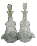 Conjunto Galheteiro Duplo Antigo em Vidro Prensado (Cruet Set), composto por duas garrafas com rolhas de vidro e base unificada. Padrão de folhagem/ramagem gravado na ampulheta, com base prensada com estrela e alças decoradas. Marcas do tempo. -dim- base 18x8 garrafa 7d 17a