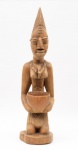 AFRICA OCIDENTAL - Estatueta africana em madeira clara, retratando figura tribal feminina com rebuscado penteado e escarificações faciais. Apresenta-se com recipiente nas mãos, ajoelhada sobre base circular. África Ocidental. OBS: (Rachaduras e marcas do tempo). Med. Alt. 52,0 cm.