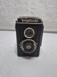 Câmera vintage Voigtländer Brillant, modelo de médio formato produzido na Alemanha nas décadas de 19301950. Corpo em metal e baquelite com detalhes cromados e lente dupla típica das TLR (Twin Lens Reflex). Acompanha estojo original em couro (No estado). Item de colecionador, com sinais do tempo, necessita de reparos.