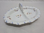 Linda petisqueira em porcelana alemã Württemberg, início do séc. XX, com formato oval bipartido, alça central modelada, bordas frisadas com detalhes em azul, decoração floral policromada pintada à mão. Medindo 24cm x 15cm.