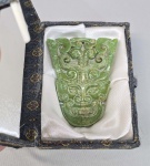 Pingente chinês esculpido em Jade verde, representando máscara no estilo arcáico. No estojo original. Med. 8 x 6 cm.