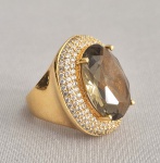 Grande anel dourado, cravejado com zircônias e grande pedra fumê central, lapidação oval. Aro 20