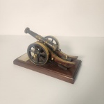Elegante miniatura de canhão com estrutura metálica em acabamento escuro e detalhes dourados, apoiada sobre sólida base de madeira polida. Rica em detalhes, a peça evoca as tradições militares e o universo naval. Pertenceu a um almirante da Marinha Brasileira, acrescentando um contexto histórico discreto ao objeto.                                                       Estado de conservação: apresenta leves sinais do tempo e pequenas marcas de manuseio, mantendo excelente aparência geral. Medindo: 13x22x12