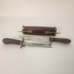 Conjunto composto por faca e garfo trinchante com cabos em madeira, acompanhado de estojo entalhado com apliques em metal. Apresenta design clássico e funcional, ideal para o preparo e serviço de carnes.                                                                                                                      Estado de conservação: bem preservado, com marcas do tempo na lâmina. Medindo: 7x45x9