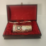 Conjunto composto por cuia em metal ricamente decorada em relevo, acompanhada de bomba metálica, acondicionado em estojo de madeira com interior revestido em tecido vermelho. A cuia apresenta acabamento trabalhado com motivos ornamentais e detalhe esmaltado em cores, conferindo caráter cerimonial e comemorativo à peça.                    Estado de conservação: bom estado geral, com sinais naturais do tempo no estojo e leve pátina no metal, sem comprometer a integridade do conjunto. Medindo: 11x27x15O