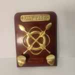 Cabideiro de parede em madeira com aplicações em Bronze representando timão e remos cruzados, encimado pela inscrição Captain. Peça de inspiração naval, com estética clássica bastante associada a ambientes marítimos e decorativos.                                                          Estado de conservação: bom estado geral, com marcas leves de uso e pátina natural nos metais. Medindo: 24x19