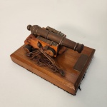 Miniatura de canhão em bronze montada sobre base de madeira, rica em detalhes construtivos que remetem à artilharia clássica. Peça de caráter ornamental, ideal para compor escritórios, bibliotecas, salas temáticas ou coleções voltadas à militaria.              Estado de conservação: Bom estado geral, com leves sinais do tempo compatíveis com a idade e a natureza decorativa da peça. Medindo: 8x14x10
