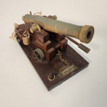 Miniatura de canhão decorativo em bronze, montado sobre base de madeira. A peça apresenta detalhes que remetem à artilharia clássica.                                                                                            Estado de conservação: Bom estado geral, com leves sinais do tempo compatíveis com a idade e a natureza decorativa da peça. Medindo: 11x19x10