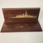 Peça institucional náutica composta por base em madeira maciça com aplicação metálica representando embarcação. A presença de marca retangular na base indica que originalmente sustentava uma placa comemorativa ou de identificação, característica comum em homenagens ou reconhecimentos no meio naval.                                                     Estado de conservação: Bom estado geral, com sinais leves de uso e marca visível onde havia a placa original, sem comprometer a integridade da peça. Medindo: 9x19x8