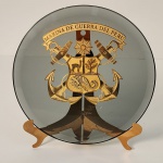 Descrição: Prato em vidro fumê com aplicação central dourada trazendo o brasão oficial da Marina de Guerra del Perú. O emblema é extremamente detalhado, composto por âncoras cruzadas, canhões, cordas navais e um escudo dividido com símbolos tradicionais do Peru, coroado por uma estrela radiante. Medindo: 22x22
