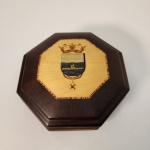 Elegante caixa octogonal em madeira nobre, com acabamento polido e interior revestido em veludo azul profundo, ideal para guardar joias ou pequenos objetos de valor. Na tampa, destaca-se um brasão trabalhado em marchetaria, representando paisagem marítima com embarcação, coroado por elementos heráldicos finamente detalhados. Medindo: 8x19x19