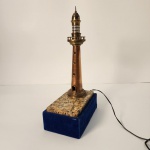 Luminária ornamental inspirada em faróis costeiros, construída em bronze. O corpo é esguio e bem trabalhado, com janelas na parte superior que simulam a área de iluminação do farol. A base combina pedra polida e revestimento aveludado azul, criando um contraste sofisticado e muito decorativo. Medindo: 39x13x20