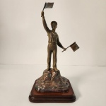 Elegante escultura em bronze representando um marinheiro em posição de sinalização, segurando duas bandeiras como se orientasse navios à distância. O metal apresenta pátina natural, valorizando os detalhes do uniforme e da base rochosa.