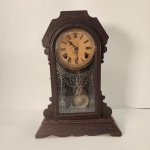 Relógio antigo confeccionado em madeira escura, com mostrador de algarismos romanos e mecanismo mecânico com pêndulo. Possui porta frontal em vidro decorado e detalhes entalhados que reforçam seu estilo clássico. Medindo: 49x33x13