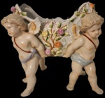 Elegante centro de mesa em porcelana Meissen, marcado na base com as tradicionais espadas cruzadas. A peça apresenta quatro querubins finamente modelados sustentando uma cesta vazada, ricamente adornada com flores aplicadas à mão e detalhes em relevo.O trabalho revela alto nível de precisão artística, com acabamento delicado, expressão suave das figuras e policromia harmoniosa. Medindo: 25x25x24