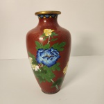 Vaso cloisonné com fundo vermelho craquelado e decoração floral policromada, destacando flores em azul, amarelo e branco com folhagens verdes. Possui formato clássico, acabamento dourado no gargalo e na base, e pintura detalhada que valoriza a peça. Medindo: 27x14