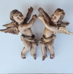 Par de putti em madeira entalhada, policromados e com douração em detalhes, representados em posição ascensional, com drapeados esvoaçantes e asas abertas trabalhadas em finas camadas. Expressões delicadas e volumetria bem modelada, com acabamento envelhecido que realça a textura da madeira e a pátina original. Peças de grande apelo decorativo, ideais para composição em parede ou em ambientes de linguagem clássica sacra. Excelente presença escultórica. 55x23x43 centímetros (cada).