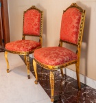 Par de cadeiras em madeira entalhada e dourada, com trabalho minucioso em folhas de acanto, laços e frisos geométricos, características associadas ao estilo Louis XVI. Molduras com pátina dourada original, apresentando desgaste natural que reforça o caráter decorativo e a autenticidade do conjunto. Encostos em formato levemente arqueado, com coroamento decorado por laço entalhado. Assentos amplos e confortáveis, revestidos em tecido brocado vermelho de padrão floral clássico. Pernas dianteiras curvas (cabriolet) e traseiras levemente arqueadas, ambas com entalhes decorativos.
