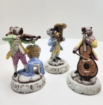 Conjunto de três figuras Capodimonte, peças finamente modeladas em porcelana com músicos de feições caninas em trajes rococó. A pintura é precisa e vibrante, com douração sutil e bases ornamentadas que reforçam a qualidade do trabalho. As poses são equilibradas, os instrumentos bem definidos e o acabamento detalhado confere presença marcante e estética refinada ao conjunto. Dimensões:: 17x17 centímetros cada.