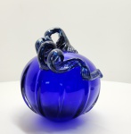 Elegante escultura em vidro artístico soprado, em formato de abóbora estilizada, de corpo facetado e tom azul cobalto profundo, com aplicação superior em vidro retorcido de tonalidade azul escuro translúcido. A peça revela domínio técnico excepcional na manipulação do vidro ainda em fusão, evidenciado na simetria das gomos, no brilho intenso e na pureza do vidro. O contraste entre o corpo liso e a alça retorcida cria um efeito visual vibrante, típico das criações modernas inspiradas na escola Murano, onde o vidro é simultaneamente matéria escultórica e luminosa. O azul cobalto uma das cores mais difíceis de obter com transparência e uniformidade é aqui explorado em toda sua profundidade, resultando em uma obra de grande presença visual e sofisticação cromática.Dimensões:20 x 16 centímetros.