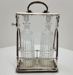 Sofisticado tantalus (porte-liqueur) de fabricação WMF Württembergische Metallwarenfabrik, Alemanha, c. 1900, composto por duas garrafas em cristal lapidado abrigadas em uma estrutura metálica prateada de elegante design geométrico. A armação, de forma trapezoidal, apresenta alça superior decorada em torção, pés esféricos e o característico mecanismo lateral com chave funcional, que permite travar as garrafas, garantindo segurança e exclusividade uma das marcas dos modelos de prestígio da WMF.As garrafas, em cristal facetado de alta pureza, exibem lapidação em faixas verticais e tampas facetadas, em perfeita harmonia com o brilho acetinado do metal prateado. Na base, punções WMF gravadas, incluindo o losango com avestruz (símbolo da manufatura) e o código I/O, indicando prata eletrolítica de alta qualidade. Dimensões: ( 38 x 23 x 13 centímetros)