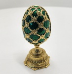 Elegante ovo decorativo no clássico estilo Fabergé, adquirido na Rússia e confeccionado em liga metálica de alta qualidade, banhada a ouro. O exterior é revestido por esmalte translúcido em tom verde esmeralda, aplicado sobre fundo texturizado em padrão guilloché, que cria profundidade, brilho e movimento característicos das peças inspiradas na tradição imperial russa. A decoração externa apresenta treliças douradas em relevo, ornadas com pequenos cristais e arabescos minuciosamente modelados. Abertura articulada em duas metades revela interior também esmaltado em esmeralda profunda, com acabamento floral em relevo. No centro destaca-se a surpresa do ovo: um mini relógio ornamental, com tampa esmaltada e estrutura banhada a ouro, seguindo o estilo dos relógios de bolso europeus. Dimensões:12x6 centímetros.